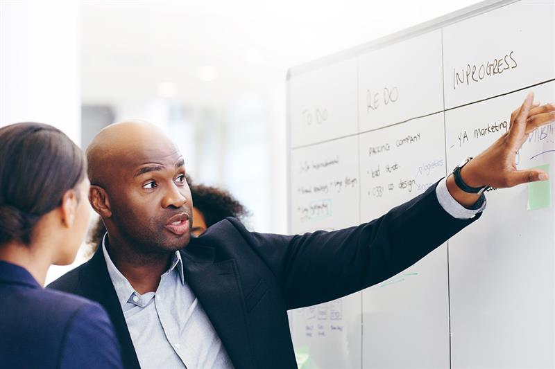 Manager explaining workflow on a whiteboard during a meeting, showcasing how LMS for employee training supports task tracking and project management.