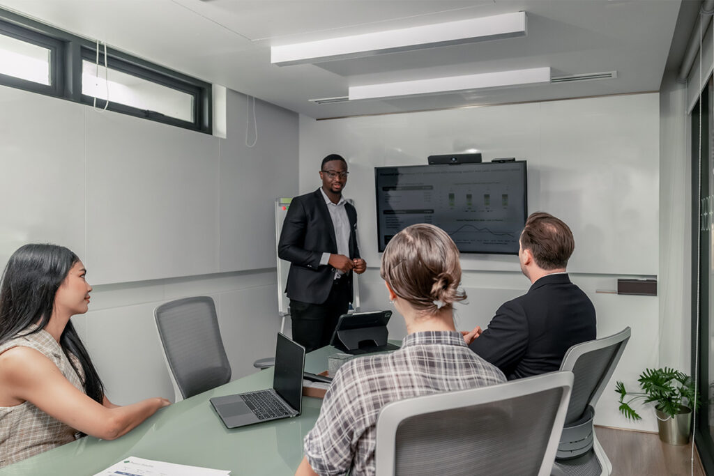 A team in a meeting room reviewing charts on a large display screen as a presenter explains data trends linked to a digital admission system for university admissions.
