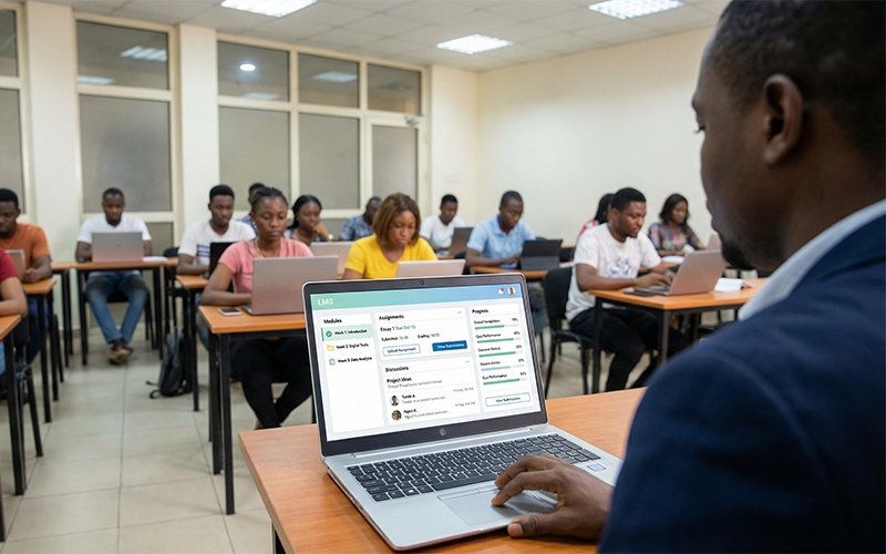 Instructor using laptop in a digital classroom environment, screen displaying LMS dashboard with: course content modules, assignment submissions, discussion forums, and student progress tracking; students visible in background using devices, blended learning setting, focus on teaching and learning workflow