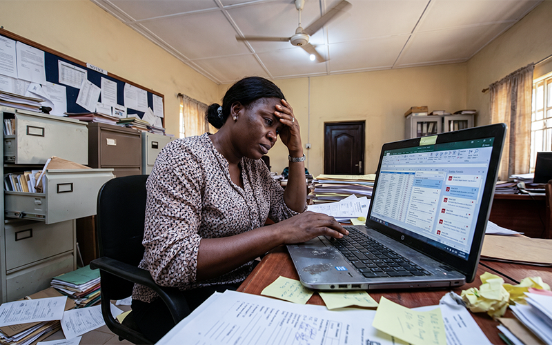 Chaotic admin desk: piles of paper applications Excel sheets open on laptop multiple emails visible staff looks overwhelmed; represents delays, manual work, and lost applicants